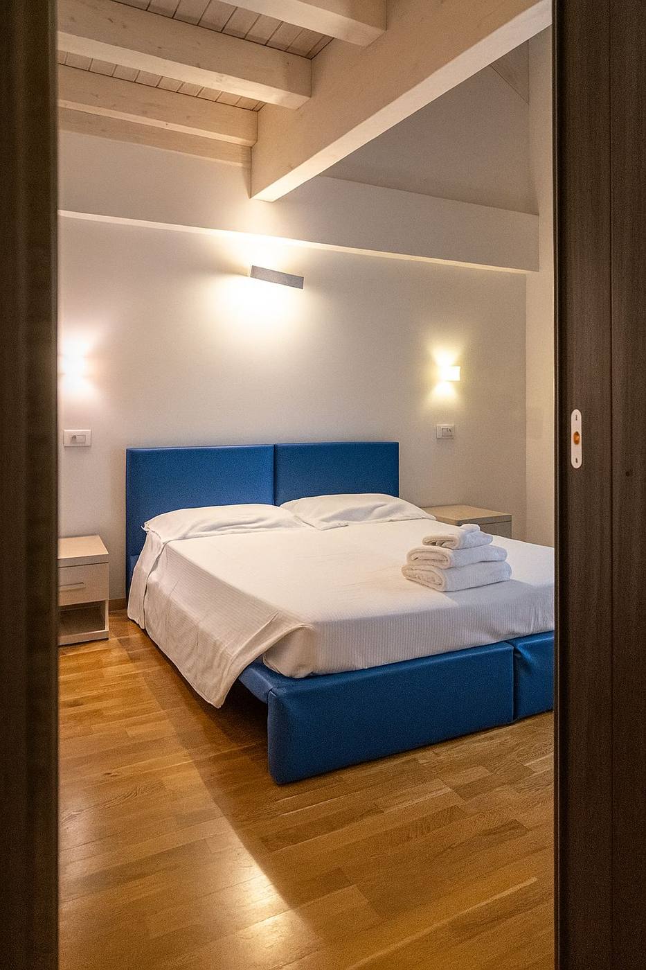 Il letto con la testiera blu sotto le travi chiare del soppalco, nella Doppia Confort Duplex di ASC Hotel